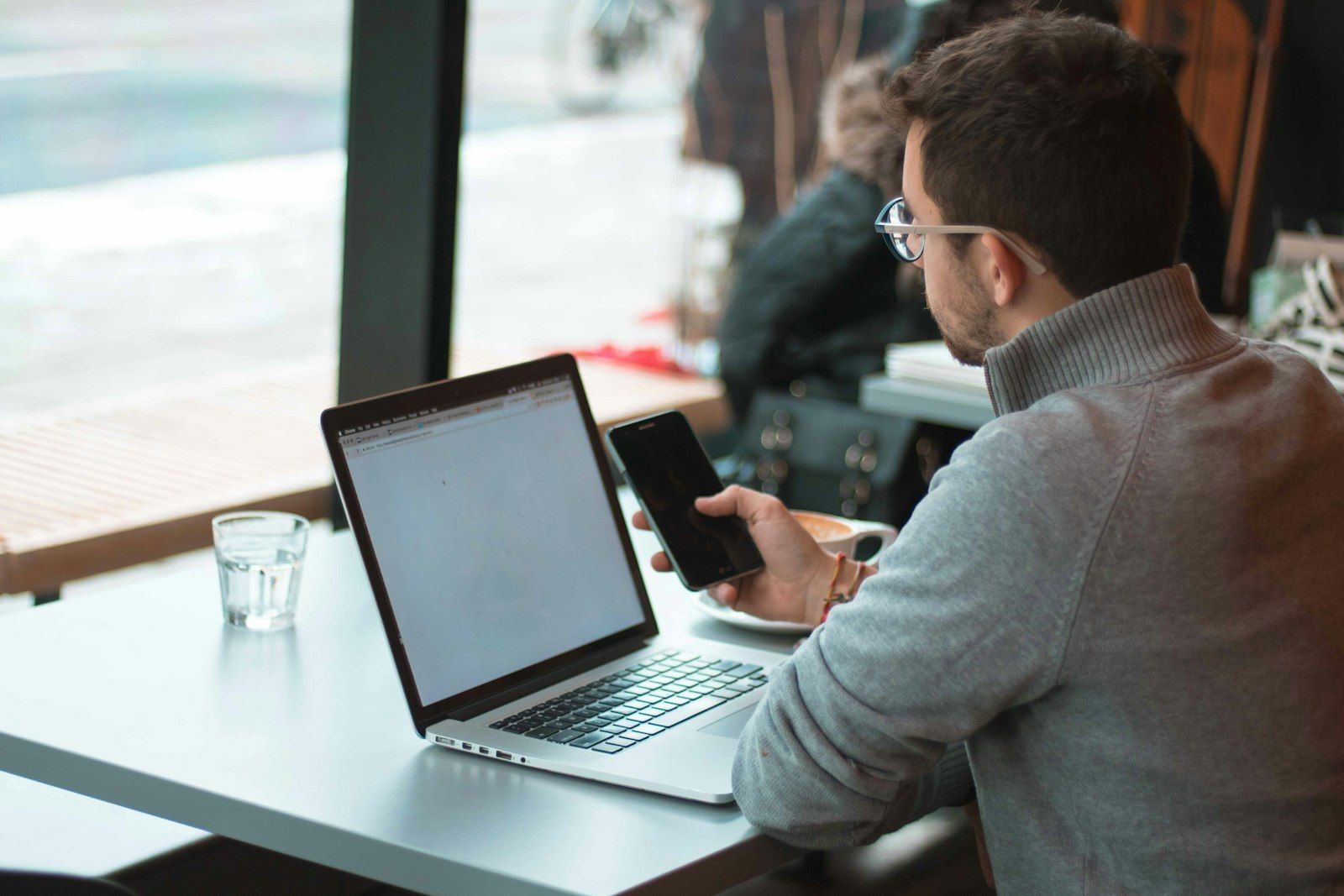 Een man achter zijn laptop die bij het raam op zijn telefoon kijkt